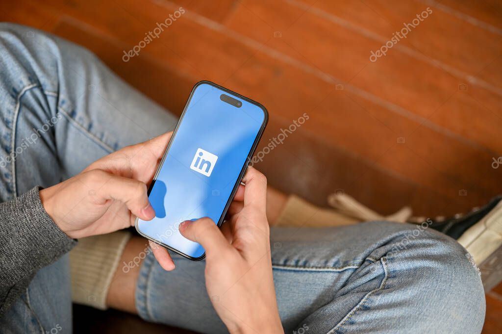 Chiang Mai, Thailand - Mar 07 2023: Top view of a man in jeans sits on the floor using LinkedIn mobile application on his phone. iPhone with LinkedIn logo on screen.