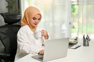 Profesyonel ve çekici Asyalı Müslüman iş kadını ya da kadın girişimci ofisindeki dizüstü bilgisayarında çalışırken kahvesini yudumluyor..