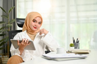 Çekici ve kendinden emin bin yıllık Asyalı Müslüman iş kadını ya da türban ya da tesettürlü kadın CEO 'su dijital tabletle masasında oturuyor ve kameraya bakıp gülümsüyor..