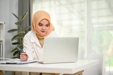 Konsantre olmuş milenyum Müslüman iş kadını masaya yaslanmış, dizüstü bilgisayar ekranına bakıyor, rapora bir şeyler not ediyor, ofisinde çalışıyor..