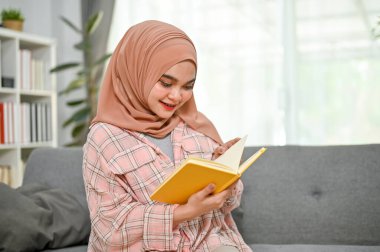 Çekici ve pozitif Asyalı Müslüman kadın gündelik giysiler içinde oturma odasında kanepede kitap okuyor..