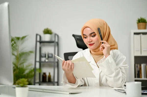 stock image Attractive and professional millennial Asian Muslim businesswoman wearing hijab reading some information on a book at her desk in the office.