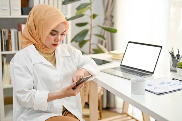 Professional and confident millennial Asian Muslim businesswoman wearing hijab using her phone at her desk in the modern private office.