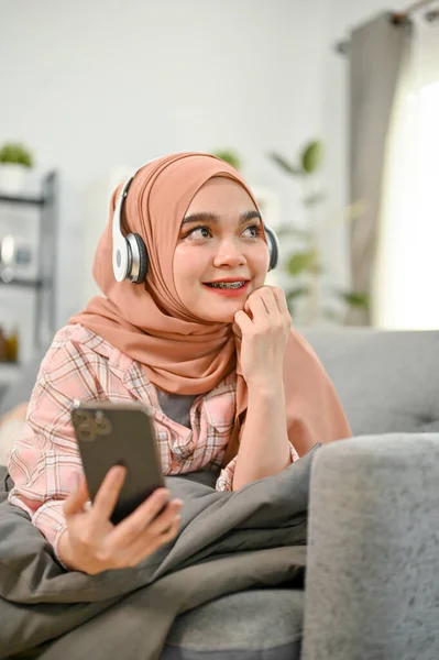 Portrait, Cheerful and pretty young Asian Muslim woman wearing hijab relaxing in her living room, listening to music through headphones on sofa.