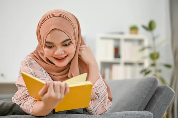 stock image Happy young Asian Muslim woman in casual clothes and hijab reading a book and laying on sofa in her living room. leisure and lifestyle concept