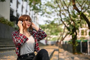 Çekici, genç, Asyalı, sıradan kıyafetler ve gözlüklerle kampüs binasının dışındaki merdivenlerde oturmuş kulaklıkla müzik dinleyen güzel bir üniversite öğrencisi..