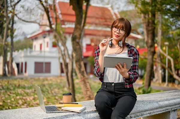 身穿法兰绒衬衫和眼镜的年轻漂亮聪明的亚洲女大学生在校园公园的长椅上 用她的数码平板电脑思考和规划她的学校项目 — 图库照片