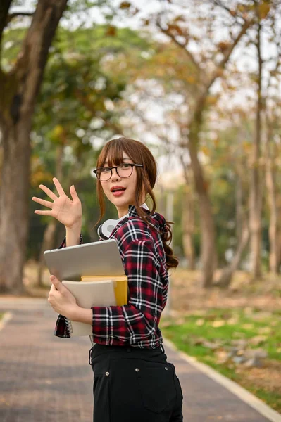 彼女のラップトップと学校の本を保持し 手を振って 挨拶ジェスチャー キャンパスパークに沿って歩くカジュアル服でかなり若いアジアの女子大学生 — ストック写真