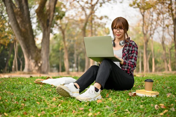 在美丽的绿树成荫的公园里 身穿休闲装的年轻漂亮的亚洲女大学生坐在草地上 一边用她的笔记本电脑 一边吸引人 — 图库照片