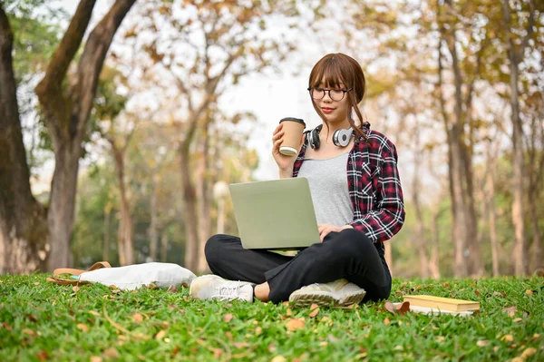 穿着休闲装的年轻亚洲女大学生坐在绿树成荫的公园的草地上 一边喝着咖啡 一边用笔记本电脑管理着自己的学业 — 图库照片