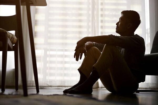 Depressed and frustrated Asian man sits on the living room floor, dark room, suffering from life failure, lack of energy, no passion, anxiety. silhouette image