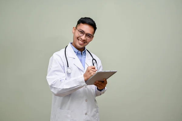 優しいと幸せな千年のアジアの男性医師で白いガウンで彼の聴診器は彼のデジタルタブレット上の医療ケースを記録します 緑のスタジオの背景に立って — ストック写真