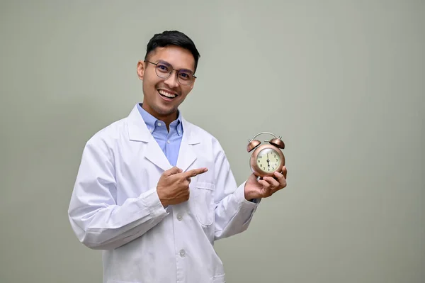 Smiling and smart millennial Asian male doctor in white gown pointing finger at an alarm clock on his hand, standing against a green studio background.