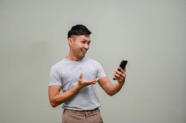 身穿休闲装的快乐快乐的千年亚洲男人看着他的手机屏幕 惊讶地发现在一个网上购物应用程序上有一个在线促销活动 这与一个绿色工作室的背景正好相反 — 图库照片