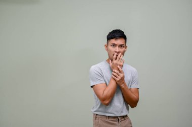 Korkan ve şok olan bin yıllık Asyalı adam, yeşil arka planda dikilirken ağzını günlük kıyafetlerle kapatıyor. gergin, korkmuş, korkmuş, korku, ürkmüş yüz ifadesi