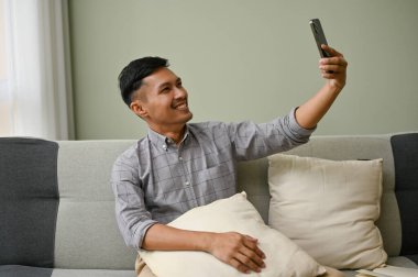 Yakışıklı ve mutlu bir Asyalı adam. Günlük giysiler içinde. Oturma odasındaki kanepede otururken telefonuyla selfie ya da video çekiyor. eğlence ve teknoloji konsepti