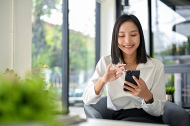Gülümseyen ve mutlu Asyalı iş kadını arkadaşlarıyla telefonda sohbet ediyor, sosyal medyada kayıyor ya da internette video izliyor, ofiste dinlenirken telefonunu kullanıyor..