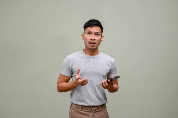 身穿休闲装的千禧年亚洲男人站在与世隔绝的绿色背景下 手里拿着手机 吓得面无血色 — 图库照片