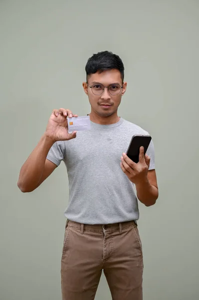 身穿休闲装的英俊的千年亚洲男人 拿着智能手机和信用卡 站在一个绿色工作室的背景下 — 图库照片