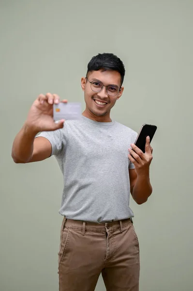身穿休闲装 拿着智能手机 站在一个绿色工作室的背景下 拿着一张信用卡拍照的快乐而微笑的千年亚洲男人 — 图库照片