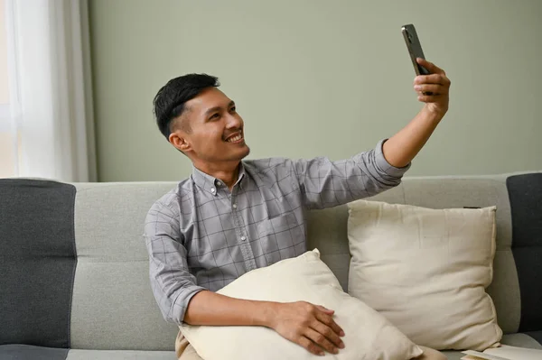 穿着休闲装的英俊而快乐的千年亚洲男人坐在客厅的沙发上 用手机自拍或录像 休闲及科技概念 — 图库照片