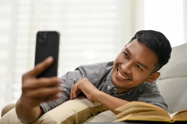 一个英俊 面带微笑的千年亚洲男人躺在客厅的沙发上 用手机自拍 周末呆在家里 — 图库照片