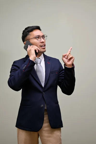 Professional millennial Asian businessman in formal business suit talking on the phone with someone, looking aside and pointing his finger up while standing against a green studio background.
