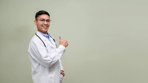 身穿白衣的快乐的千年亚洲男医生站在一个孤立的绿色背景下 站在那里展示你的文字 同时露出大拇指 微笑着 看着相机 — 图库照片