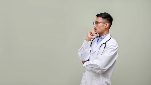 白いガウンと眼鏡で思慮深く プロの千年紀アジアの男性医師は あごに手を 空の空間を脇に見て 何かを考えて 隔離された背景 — ストック写真