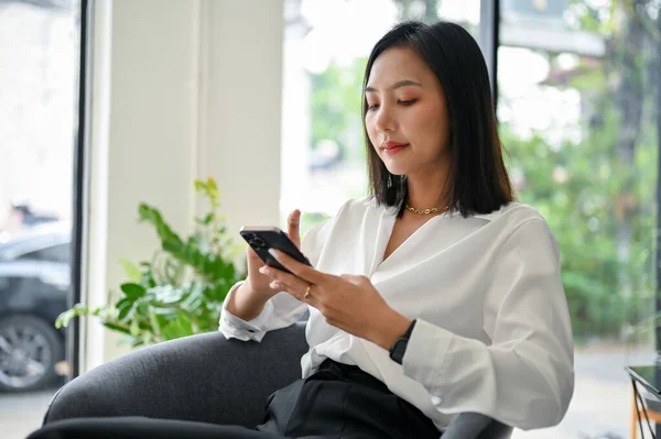 Attractive and gorgeous millennial Asian businesswoman using her phone, chatting with someone or using mobile app while relaxing in her private office.