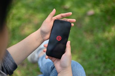 Chiang Mai, Tayland - 16 Mart 2023: Cep telefonunda Pinterest uygulaması kullanan Asyalı bir kadın. Ekranda Pinterest logosu olan iPhone. yakın plan resim