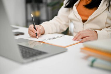 Asyalı bir üniversite öğrencisinin ev ödevi yaparken, defterine bir şeyler yazarken ve dizüstü bilgisayar kullanırken çekilmiş görüntüsü..