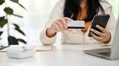 Telefonunu ve kredi kartını elinde tutan Asyalı bir kadının internet faturalarını ödemek için mobil bankacılık başvurusunu kullanırken çekilmiş görüntüsü..