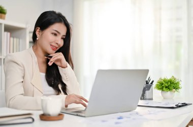 Profesyonel ve düşünceli Asyalı iş kadını dizüstü bilgisayardaki iş projesine odaklanıyor, özel ofisinde çalışıyor..