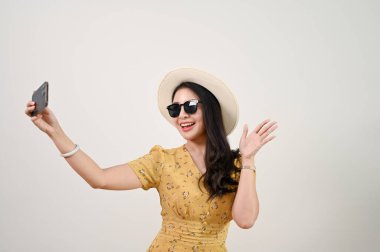 Neşeli ve neşeli bin yıllık Asyalı kadın turist hasır şapkalı ve güneş gözlüklü. Akıllı telefonuyla selfie çekerken izole edilmiş beyaz arka planda duruyor..