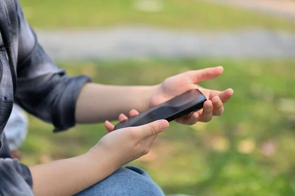 Cropped side view image of a woman in flannel shirt using her phone in the park. people and technology concept