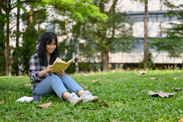 身穿休闲装的年轻漂亮的亚洲女大学生坐在草地上 在校园里看书 — 图库照片