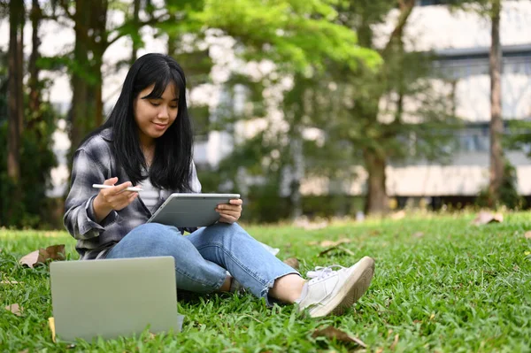 快乐的年轻亚洲女大学生穿着休闲装 坐在校园公园的草地上悠闲自在地使用她的数码平板电脑 — 图库照片