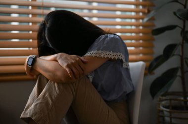 Üzgün ve depresif genç Asyalı kadın karanlık oturma odasındaki pencerenin yanında oturuyor. Zihinsel sağlık sorunu, mutsuz, yaşam yetersizliği, stres