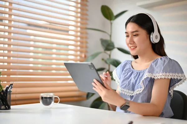 Attractive and happy young Asian woman in casual clothe using her digital tablet and listening to music through her headphones at her desk.