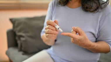 60 yaşında, hasta, emekli Asyalı bir kadının evinde kan şekerini ölçmek için parmağına mızrak taktığı yakın plan bir görüntü. Diyabet, glisemik ve sağlık hizmetleri kavramı.