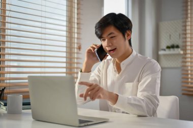 Başarılı ve profesyonel Asyalı iş adamı veya erkek yöneticinin iş ortağıyla resmi bir telefon görüşmesi yapması, dizüstü bilgisayarından bazı detaylara bakması ve ofisinde çalışması..