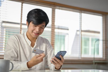 Mutlu ve yakışıklı Asyalı adam cep telefonu bankacılığı uygulamasını kullanarak para aktarıyor ya da internet faturalarını ödüyor.