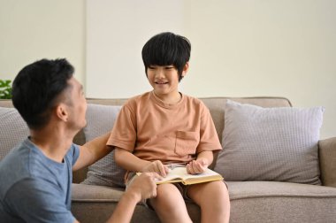 Sevimli ve mutlu bir Asyalı çocuk babasıyla konuşurken oturma odasında birlikte masal okuyor. bekar baba ve çocukluk kavramı