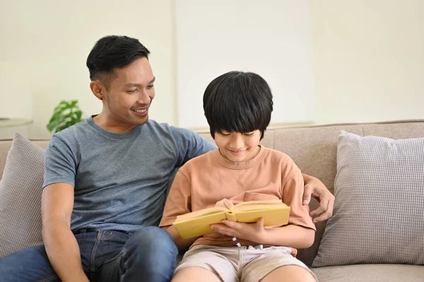Kind and caring Asian dad and his son are sitting on a comfy couch in the living room, reading a book together. family, fatherhood concept
