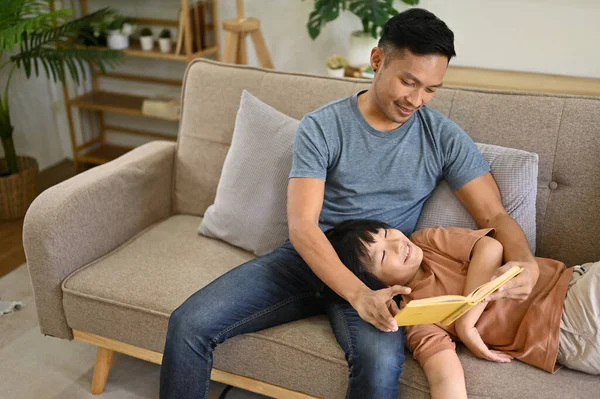 Caring and kind Asian dad telling story or reading a book with his son on sofa in the living room. happy family, parenting, single dad, fatherhood concept