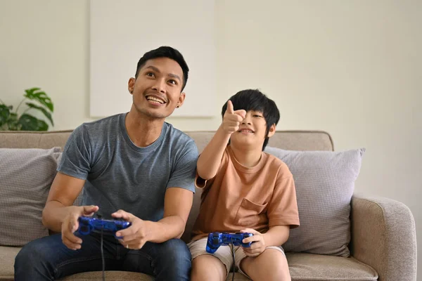 Neşeli ve heyecanlı Asyalı baba ve oğlu joystickli video oyunları oynuyorlar, online çocuk oyunlarında yarışıyorlar ve evde birlikte eğleniyorlar..