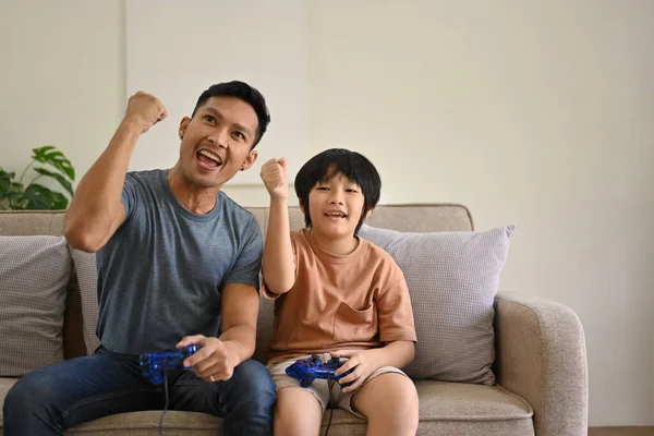 Joyful Asian dad and son with joysticks are playing video games, competing in online kid\'s games, and spending fun time at home together. happy family concept