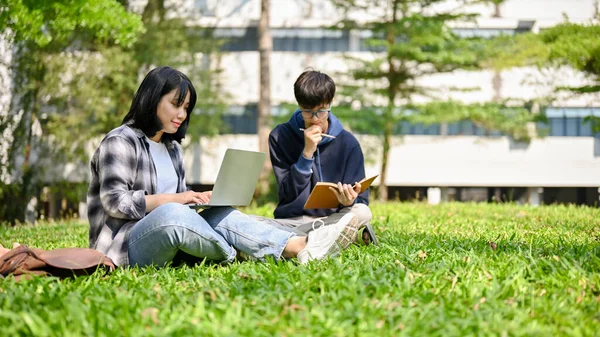 迷人而快乐的年轻亚洲女大学生与她的朋友坐在校园公园的草地上 一边用笔记本电脑一边做功课 — 图库照片