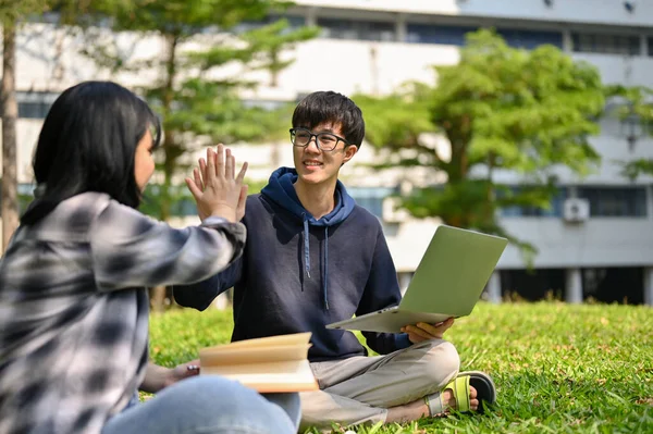 快乐的年轻亚洲男大学生一边做功课 一边坐在校园公园的草地上 一边给他的女朋友高高的五块钱 — 图库照片